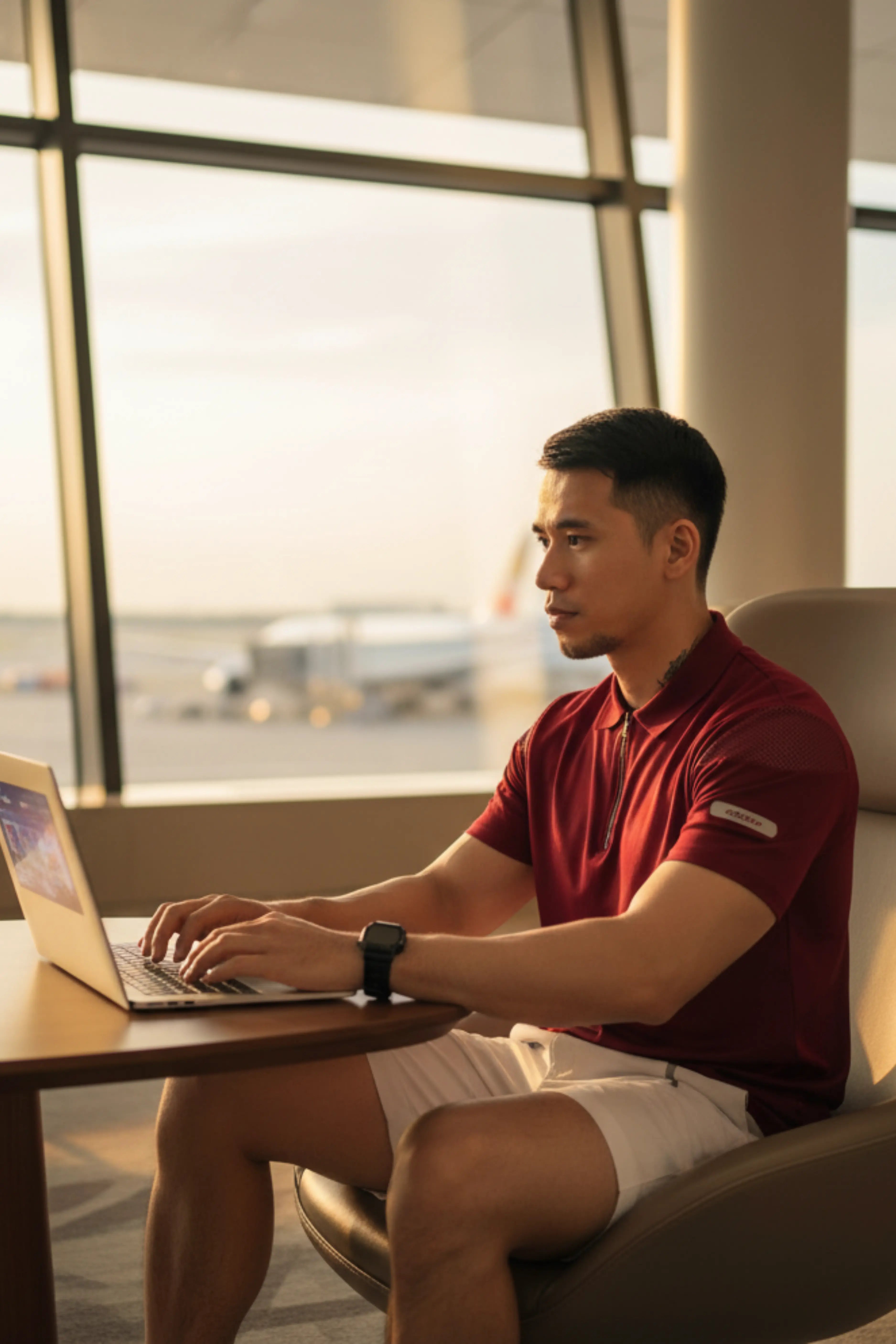 A man wearing a red polo T-Shirt at the airport typing on his laptop